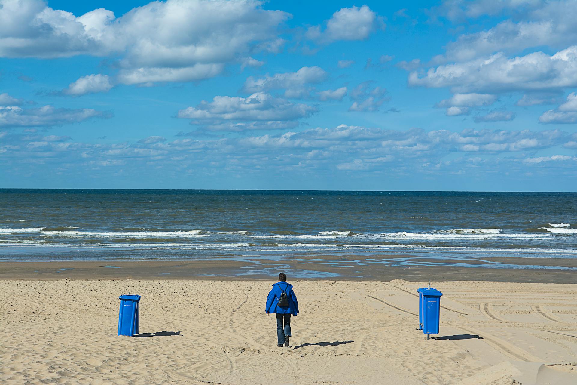 man-in-blue-jacket-in-a-beach-near-blue-trash-bins